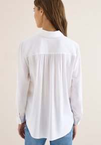Femme aux longs cheveux bruns portant une chemise blanche à manches longues boutonnée et un jean bleu, vue de dos sur un fond uni.