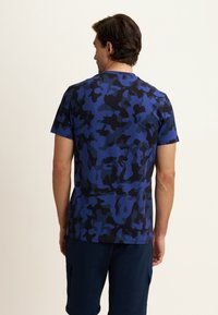 Uomo di spalle che indossa una t-shirt camouflage blu e nera con pantaloni corti blu scuro su uno sfondo chiaro e uniforme.