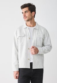 Cămașă cu butoni de culoare gri deschis, fabricată dintr-un material moale, având două buzunare pe piept, guler și o croială lejeră, purtată peste un tricou alb.