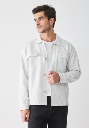 Ljust grå skjorta med knappar i mjukt material, har två bröstfickor, krage och en avslappnad passform, bärs över en vit t-shirt.