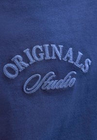 Tejido azul marino con bordado en relieve de color azul claro que contiene las palabras "ORIGINALS Studio" dispuestas en una curva. Textura suave.