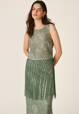 Mujer con cabello largo y oscuro que lleva un vestido verde sin mangas de crochet con patrones florales y detalles de flecos largos en la cintura.