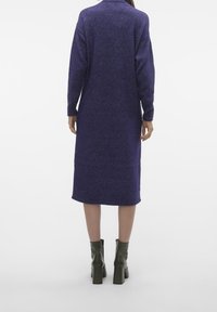 Abito maglione viola lavorato a maglia con lunghezza a metà polpaccio, maniche lunghe e colletto. Abbinato a stivaletti alla caviglia con tacco verde. Tessuto strutturato.
