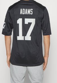 En person iført en sort sportsjersey med navnet "ADAMS" og nummeret "17" i hvidt på ryggen, parret med lysegrå bukser.