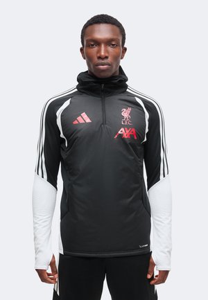 Jeune homme portant un haut d’entraînement Liverpool FC noir et blanc avec des logos rouges, debout face à un fond uni.