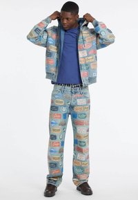 Mężczyzna ubrany w dwuczęściowy, dżinsowy strój z kolorowymi, vintage-stylizowanymi naszywkami, niebieską koszulę i ciemnobrązowe buty, poprawiający kołnierz kurtki.