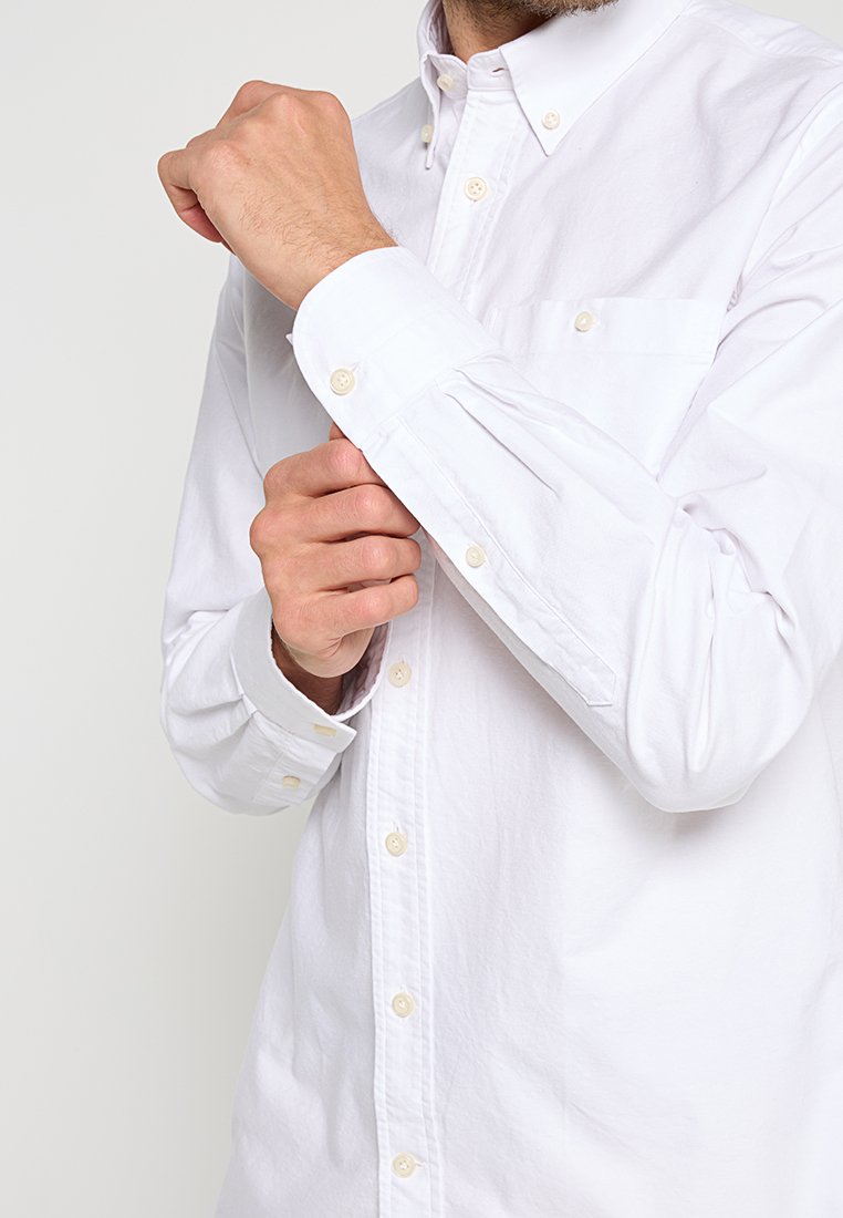 Man die de knoop van de manchet van een langemouw wit overhemd met borstzak vastmaakt tegen een effen achtergrond.