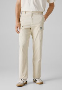 Beige cargobroek met een gestructureerde oppervlakte, zijzakken en een labelpatch op de rechterdij, gedragen met casual sneakers.