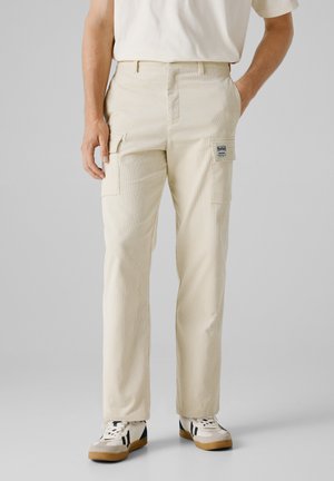 Beige Cargo-Hosen mit strukturiertem Oberflächen, Seitentaschen und einem Patcheslabel am rechten Oberschenkel, kombiniert mit lässigen Sneakers.