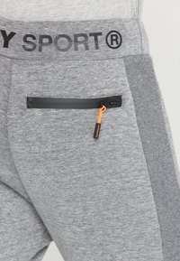 Grijze sportbroek van een gestructureerde stof met een zwarte tailleband en een zijzak met ritssluiting, verfraaid met een oranje trekkin.