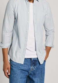 Jasnoniebieska koszula w paski z białymi i brzoskwiniowymi paskami, z długimi rękawami podwiniętymi, noszona na białym t-shircie i niebieskich dżinsach.
