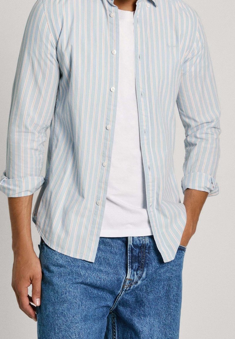Jasnoniebieska koszula w paski z białymi i brzoskwiniowymi paskami, z długimi rękawami podwiniętymi, noszona na białym t-shircie i niebieskich dżinsach.