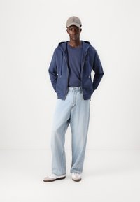 Marineblå zip-up hættetrøje over en marineblå skjorte, parret med lyseblå løse jeans og en grå kasket. Hvide sneakers med brune accenter.