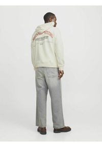 Kremfärgad hoodie med text och logotyp bak, i kombination med ljusgrå vida jeans och bruna skor, mot en enfärgad bakgrund.