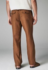 Pantalons en lin marron avec des jambes droites, dotés de deux poches arrière et de fermetures à boutons. Texture lisse, design décontracté.