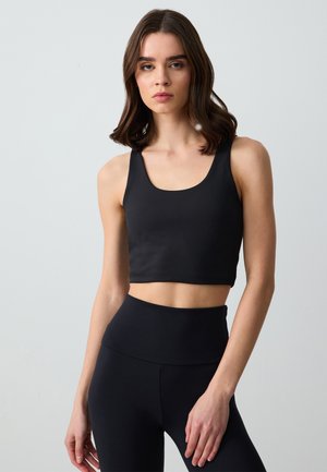 Zwarte crop top van glad, rekbaar materiaal, met een ronde halslijn en brede bandjes. Gelaagd met zwarte high-waisted leggings.