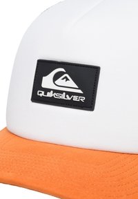 Hvid kasket med en orange skygge, prydet med et sort rektangulært mærke med et bølge-logo og mærkenavnet "Quiksilver" i hvid skrift.