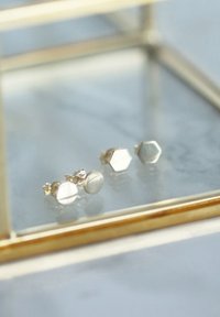 Zilveren stud oorbellen met een cirkelvorm en een hexagonale vorm, beide glad en reflecterend, tentoongesteld in een glazen vitrine met gouden accenten.