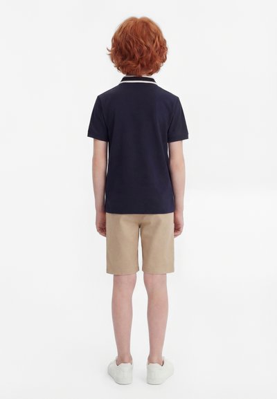 Otrok z rdečimi lasmi, oblečen v mornarsko modro polo majico s kratkimi rokavi, bež kratke hlače in bele superge, stoji obrnjeno stran na belem ozadju.