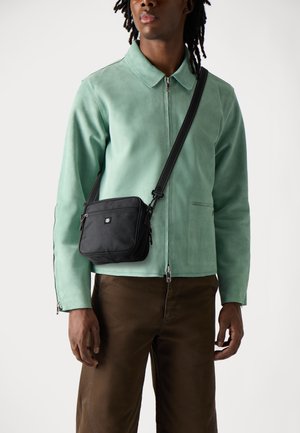 Black fabric shoulder bag with adjustable strap, zippered compartments, and a logo patch, worn over a light green suede jacket and brown pants.