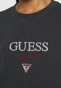 Sudadera negra con cuello redondo acanalado. Presenta el logo bordado "GUESS" en plata y "originals" en rojo, además de un detalle de logo triangular.
