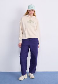 Kremfarget hettegenser med grønn logo, lilla bukser og lyseblå cap. Teksturerte sneakers fullfører antrekket, stående mot en ensfarget bakgrunn.