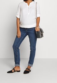 Bílá blůzka s krátkým rukávem a výstřihem ve stylu henley se třemi knoflíky. V kombinaci s modrými džíny a černými nazouvacími botami se páskem.