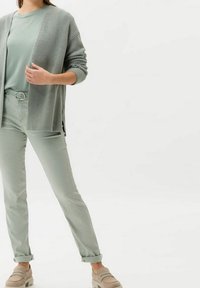Kvinna i ljusgröna högmidjade byxor, matchande t-shirt, stickad cardigan och beige loafers mot en enkel vit bakgrund.