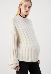 Krämfärgad stickad tröja med vertikal ribbad struktur, oversized ärmar och svart kant vid halsringning och ärmslut, kombinerad med svarta byxor.