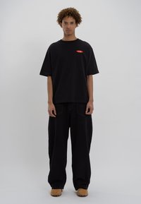 Black cotton t-shirt with short sleeves, features red logo on the chest. Paired with loose black pants and tan leather shoes. Simple design.