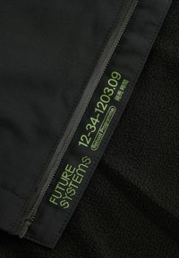 Tissu noir avec une couche extérieure lisse et une doublure intérieure texturée. Le texte vert indique "SYSTÈMES DU FUTUR" avec un détail de fermeture éclair.