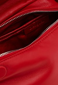 Piros bőr táska belső része cipzáros nyílással, sima felülettel és egy kis, dombornyomott címkével, amelyen a márkaneve szerepel.