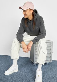 Gray hooded sweatshirt, white cargo pants with side piping, and white sneakers. Light pink cap with logo. Braided hair and sitting posture.