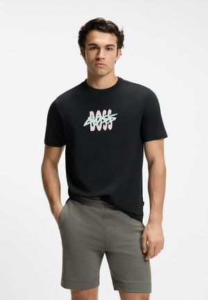 Junger Mann trägt schwarzes T-Shirt mit pastellfarbenem "BOSS"-Logo, durchgestrichen, und olivgrüne Shorts, steht mit den Händen in den Taschen.