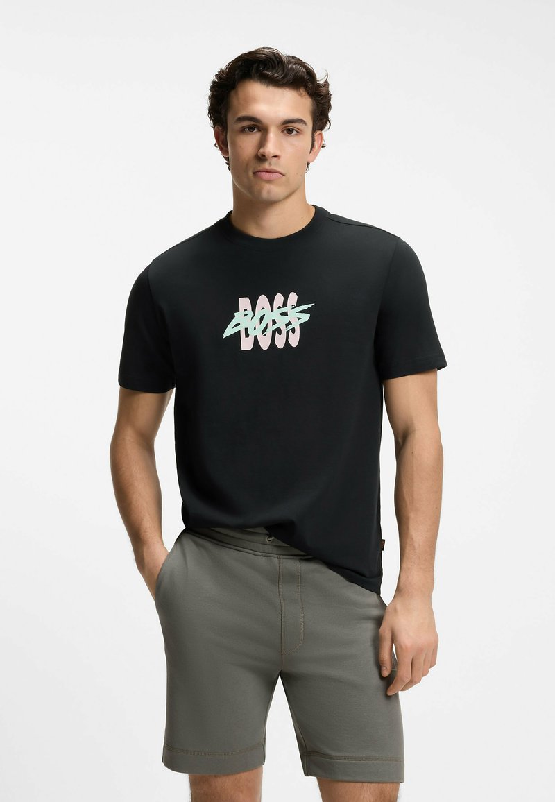 Jeune homme portant un t-shirt noir avec le logo "BOSS" pastel barré et un short vert olive, debout avec les mains dans les poches.