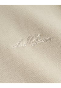 Tejido beige con un elegante logo bordado en un tono ligeramente más claro. La textura parece suave con un tejido fino.