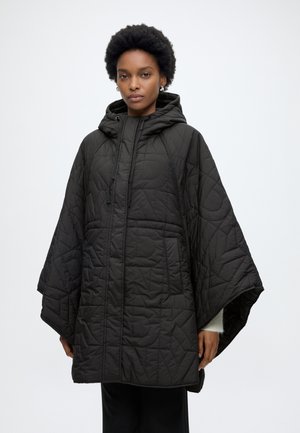 Žena nosiaca čierny prešívaný pončo s kapucňou, širokými rukávmi a prednými vreckami, stojaca na jednofarebnom bielom pozadí.