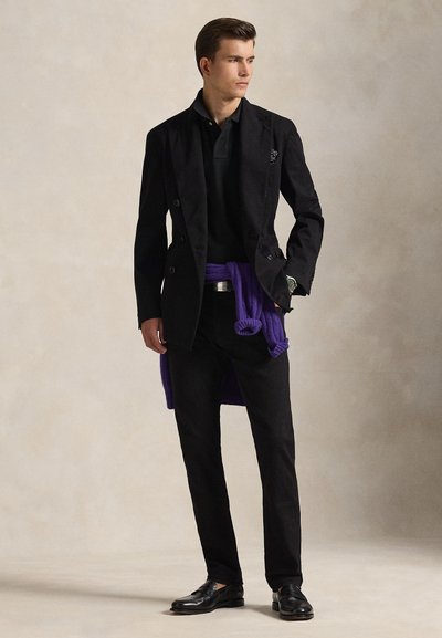 Mann trägt einen schwarzen Anzug mit schwarzem Polohemd, schwarzen Hosen, schwarzen Loafers und einem lila Pullover, der um die Taille gebunden ist, steht vor einem einfarbigen Hintergrund.