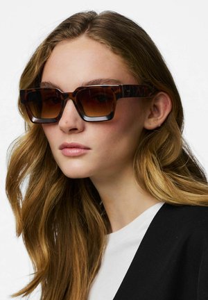 Kvinde med langt brunt hår iført store firkantede solbriller i skildpaddemønster, hvid skjorte og sort jakke mod hvid baggrund.