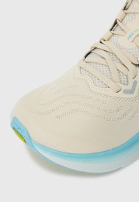 Chaussure de sport beige clair avec un dessus texturé, présentant un design en maille, une semelle bleu clair et des motifs en vagues sur le bout.