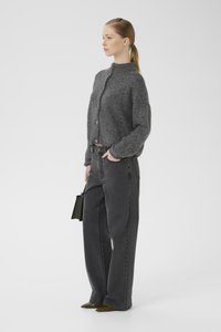 Grau strukturierte Strickjacke mit hohem Kragen und Knopfverschluss; kombiniert mit hochgeschnittenen grauen Jeans und olivgrünen spitzen Schuhen.