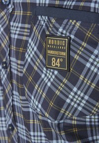 Blau- und gelbkarierter Hemd mit einer Brusttasche und einem schwarzen Etikett mit der Aufschrift "NORDIC WOODLANDS VANDERSTORM 84°". Glatte Stoffbeschaffenheit.