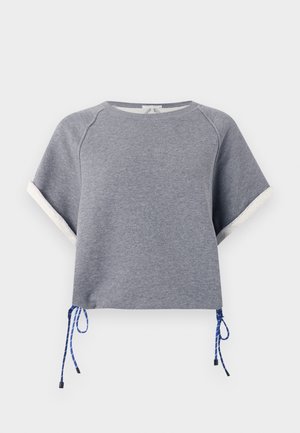 Kvinde iført en grå kortærmet sweatshirt, løstsiddende lyseblå jeans og gule åbne sandaler, stående med den ene hånd i lommen.