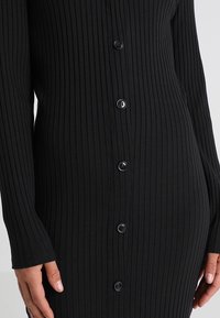 Svart rippad kl änning med långa ärmar, knappstängning fram och figurnära siluett. Texturerad tyg ökar det visuella intresset.