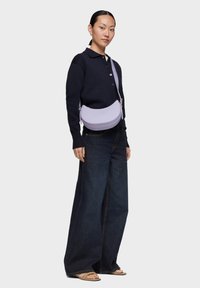 Borsa a tracolla in pelle viola con forma arrotondata e logo impresso. Il modello indossa una maglia blu navy e pantaloni denim a gamba larga. Dettagli semplici in metallo.