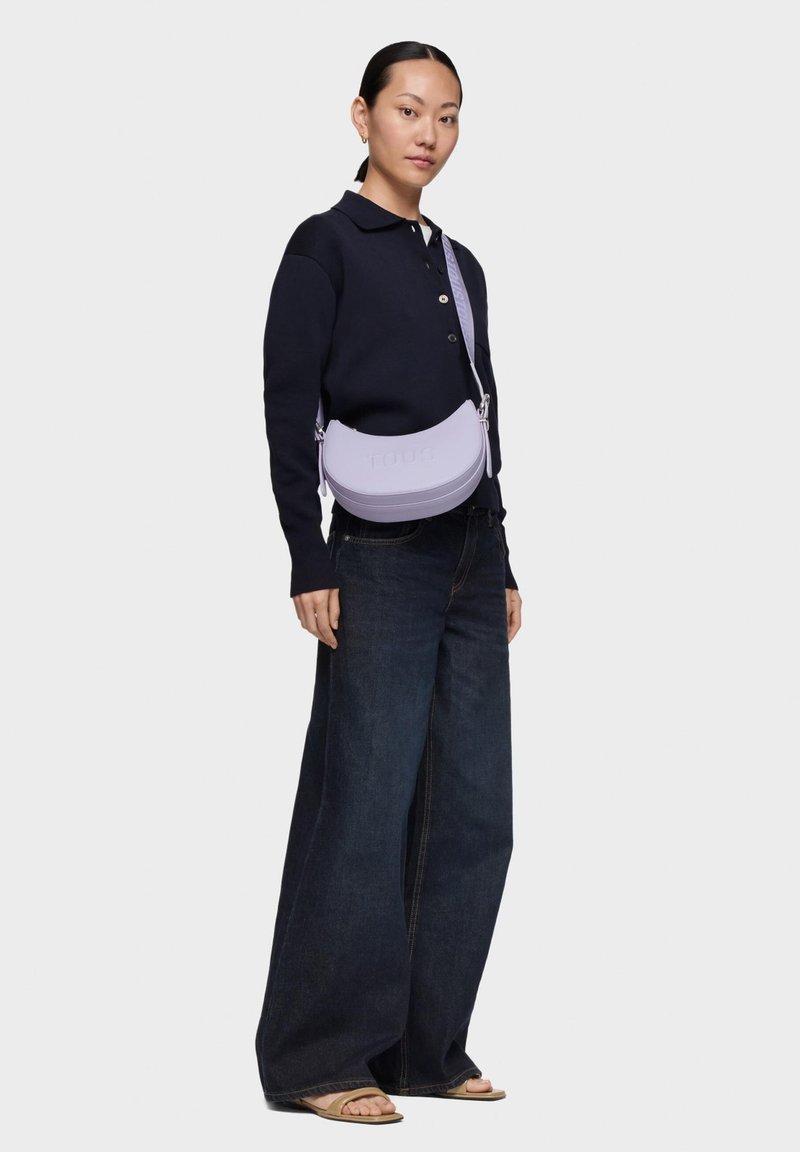 Borsa a tracolla in pelle viola con forma arrotondata e logo impresso. Il modello indossa una maglia blu navy e pantaloni denim a gamba larga. Dettagli semplici in metallo.