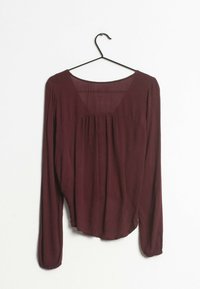 Blusa burdeos de manga larga hecha de una tela ligera, con escote redondo, frente plisado y dobladillo ligeramente curvado. Se cuelga de un gancho negro.