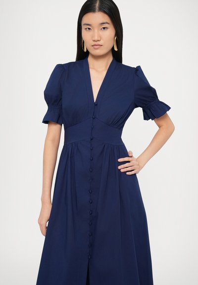 Μπλε ναυτικό φόρεμα με κοντά μανίκια-φουσκωτά, V-λαιμόκοψη και κούμπωμα με κουμπιά στο μπροστινό μέρος. Διαθέτει εφαρμοστό μπούστο και φαρδιά φούστα, κατασκευασμένο από ελαφρύ ύφασμα.