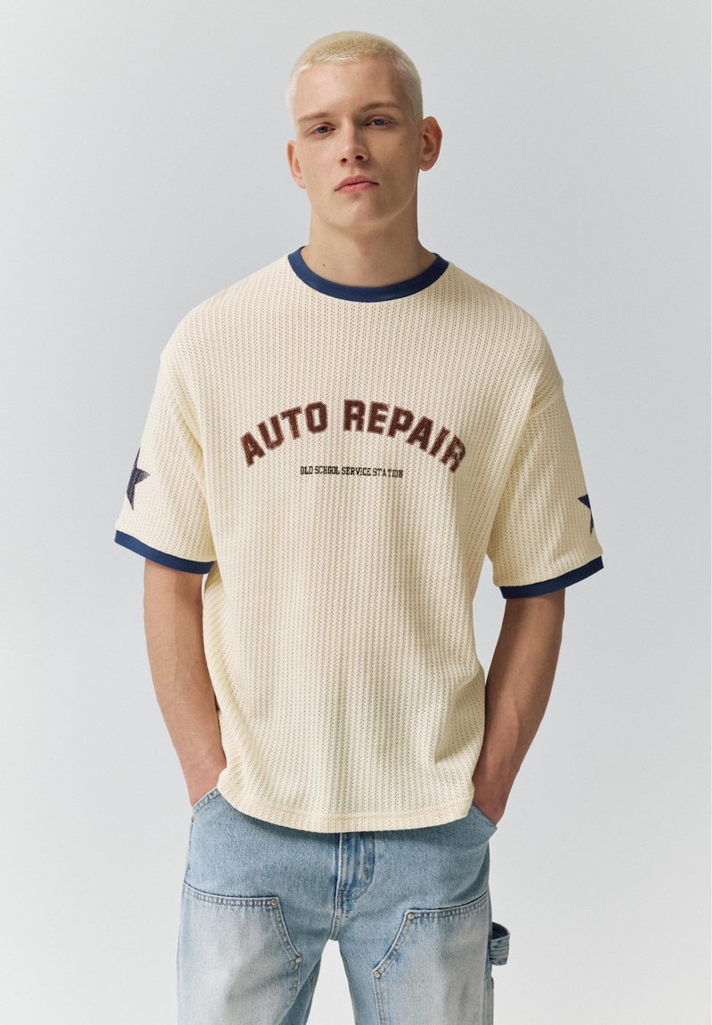 Jeune homme aux cheveux blonds courts portant un t-shirt tricoté crème avec le texte "AUTO REPAIR" et un jean bleu clair, debout les mains dans les poches.