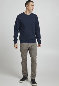 Jūras spēku sporta krekls ar apļveida kakla izgriezumu, ribotām piedurknēm un apakšmalu; apvienots ar gaiši pelēkiem biksēm un melniem sporta apaviem, stāvot pret neitrālu fonu.